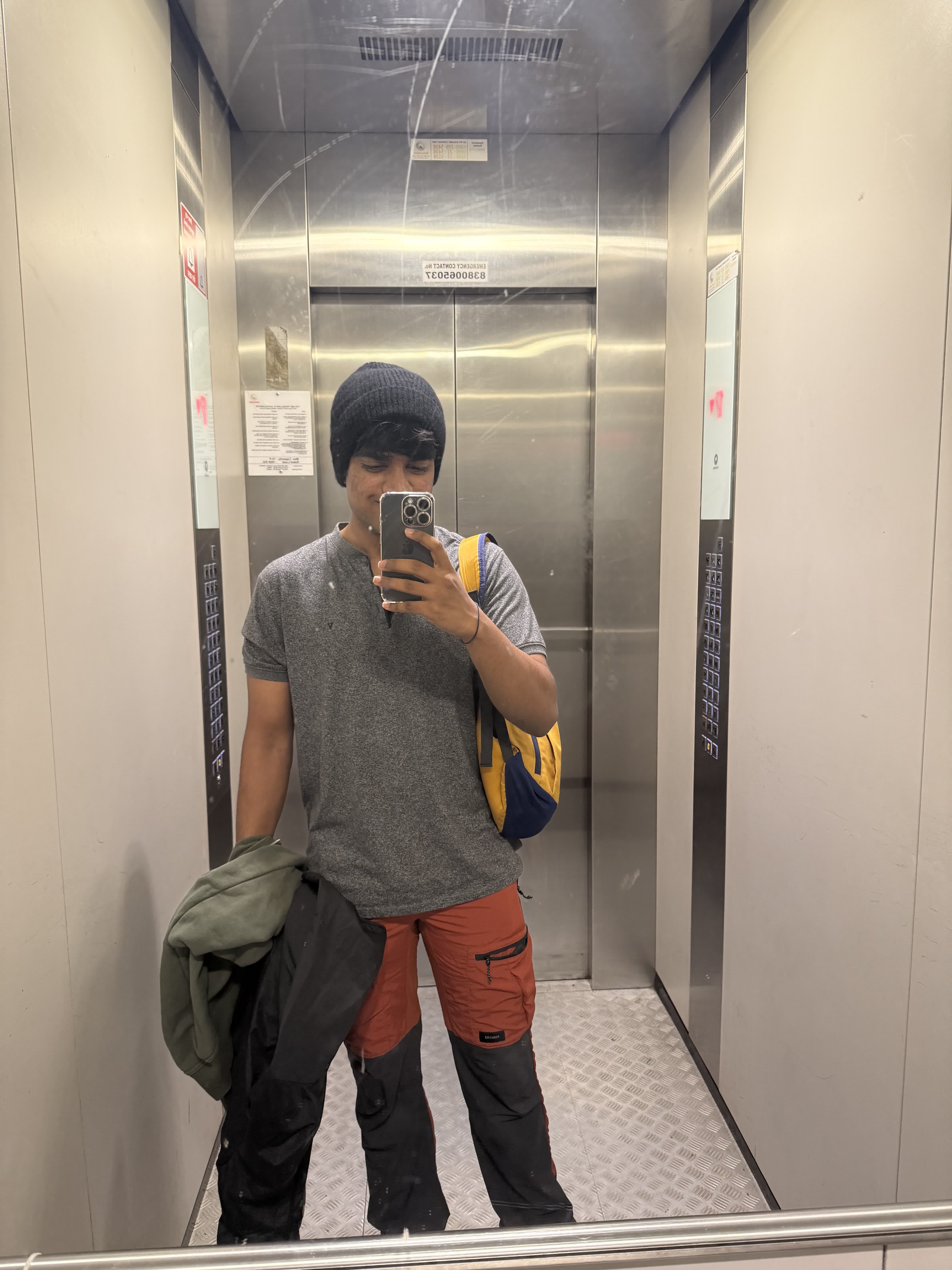 Mirror selfie in a lift, geared up with a beanie and backpack before the trek